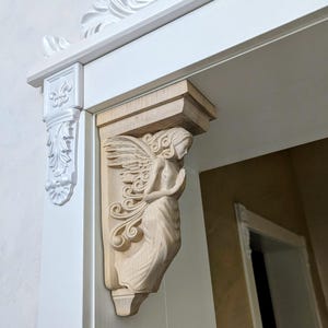 May include: A carved wooden angel corbel, with detailed wings and flowing robes, is mounted on a white door frame. The angel is in a praying pose. Decorative white molding and trim are visible.