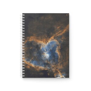 Op de afbeelding: Spiraalgebonden notitieboek met een opvallende afbeelding van een nevel. De omslag toont een hartvormige formatie in blauwe, oranje en bruine tinten tegen een donkere achtergrond, wat een kosmische esthetiek creëert. Het notitieboek heeft een witte spiraalbinding.