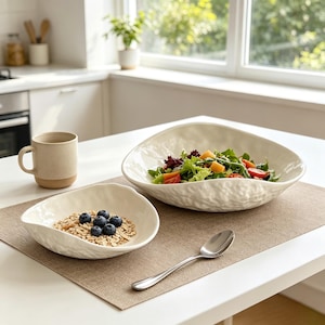 Pode incluir: Um conjunto de duas taças de cerâmica em tom esbranquiçado com uma forma orgânica irregular e uma superfície exterior texturizada. Uma taça contém uma salada verde fresca com tomates, enquanto a mais pequena contém aveia com mirtilos frescos.