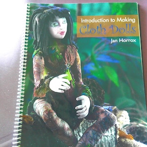May include: A spiral-bound instructional book titled Introduction to Making Cloth Dolls by Jan Horrox. The cover features a photograph of a doll with dark hair, green eyes, and patterned clothing sitting on a log against a green foliage background.