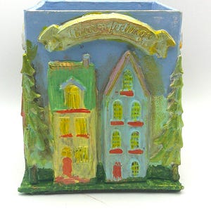May include: A square, hand-painted ceramic container with a light blue background. The container features two houses, green trees, and a banner that reads "Forever Feelings". The houses are painted in yellow, red, and blue.