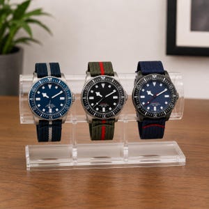May include: Three wristwatches displayed on a clear acrylic stand. The left watch has a blue dial and navy strap with a silver stripe. The center watch has a black dial and olive strap with a red stripe. The right watch has a blue dial and navy strap.
