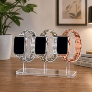 May include: A clear acrylic watch stand displaying three smartwatches with different metal bands. The watches have black screens and are held in place by clear supports. The bands are silver, gold, and rose gold.