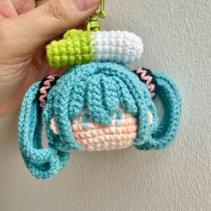 Peut inclure: Un porte-clés en crochet représentant une tête de personnage aux cheveux bleu sarcelle coiffés en deux longues couettes avec des accents noirs. Le personnage a un visage couleur pêche et est surmonté d'un petit accessoire en forme de poireau.