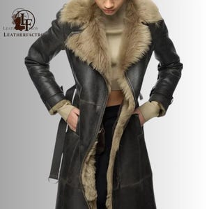 Könnte beinhalten: Ein dunkelgrauer Ledermantel mit einem Kragen und Futter aus beigefarbenem Fell. Der Mantel hat einen Gürtel, Taschen und lange Ärmel. Das Leather Factory-Logo ist sichtbar.