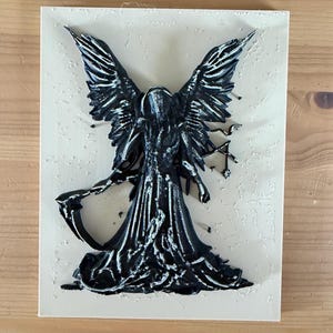 May include: A rectangular off-white plaque featuring a raised, three-dimensional black figure with large wings, a hooded cloak, and a scythe. The figure has silver-toned highlights on the edges of the wings and robes, creating a textured appearance.