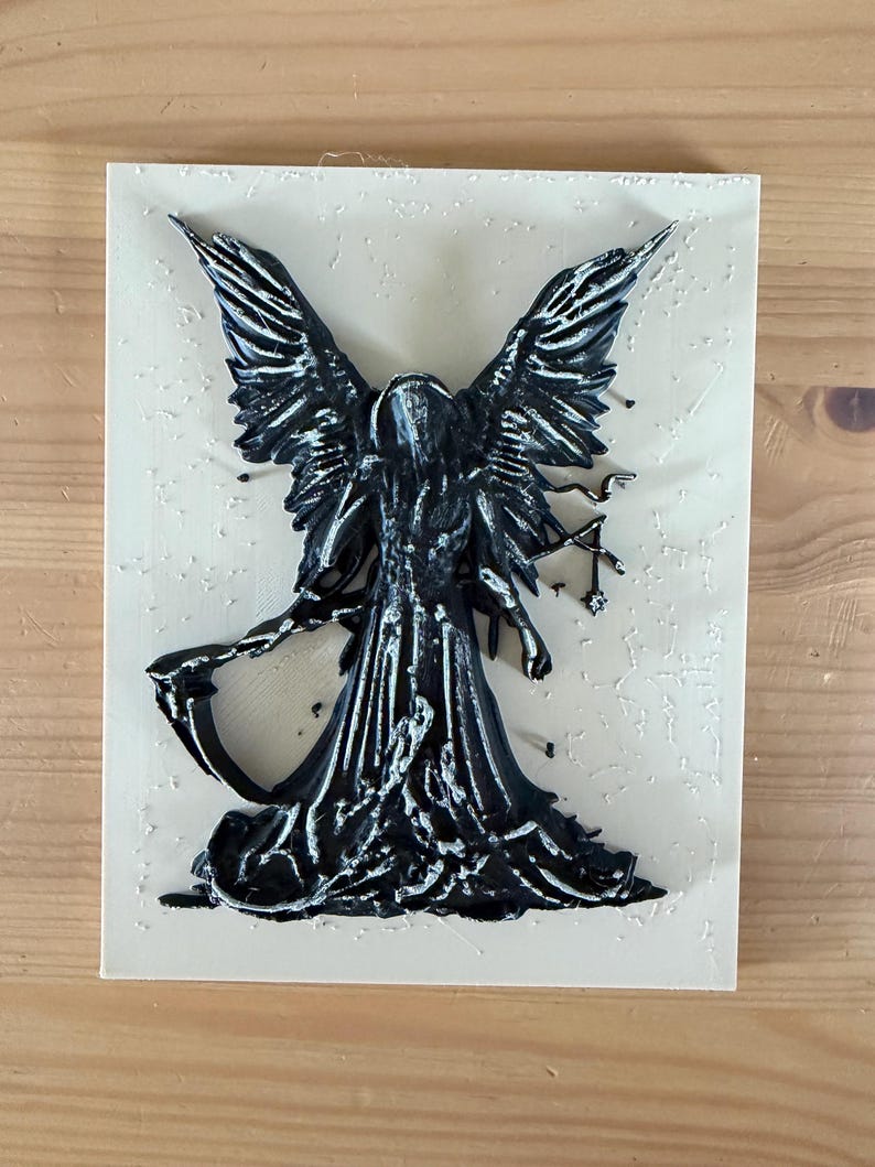May include: A rectangular off-white plaque featuring a raised, three-dimensional black figure with large wings, a hooded cloak, and a scythe. The figure has silver-toned highlights on the edges of the wings and robes, creating a textured appearance.
