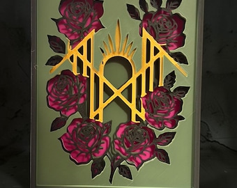 May include: A rectangular wall art piece featuring a layered design with a sage green frame. The center displays a gold-colored geometric emblem with a sunburst top. Surrounding the emblem are several deep pink roses with black outlines.