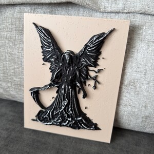 May include: A rectangular plaque with a light beige textured surface featuring a raised, three-dimensional black figure of a winged entity. The figure has large feathered wings, a hooded cloak, and holds a scythe in its right hand.