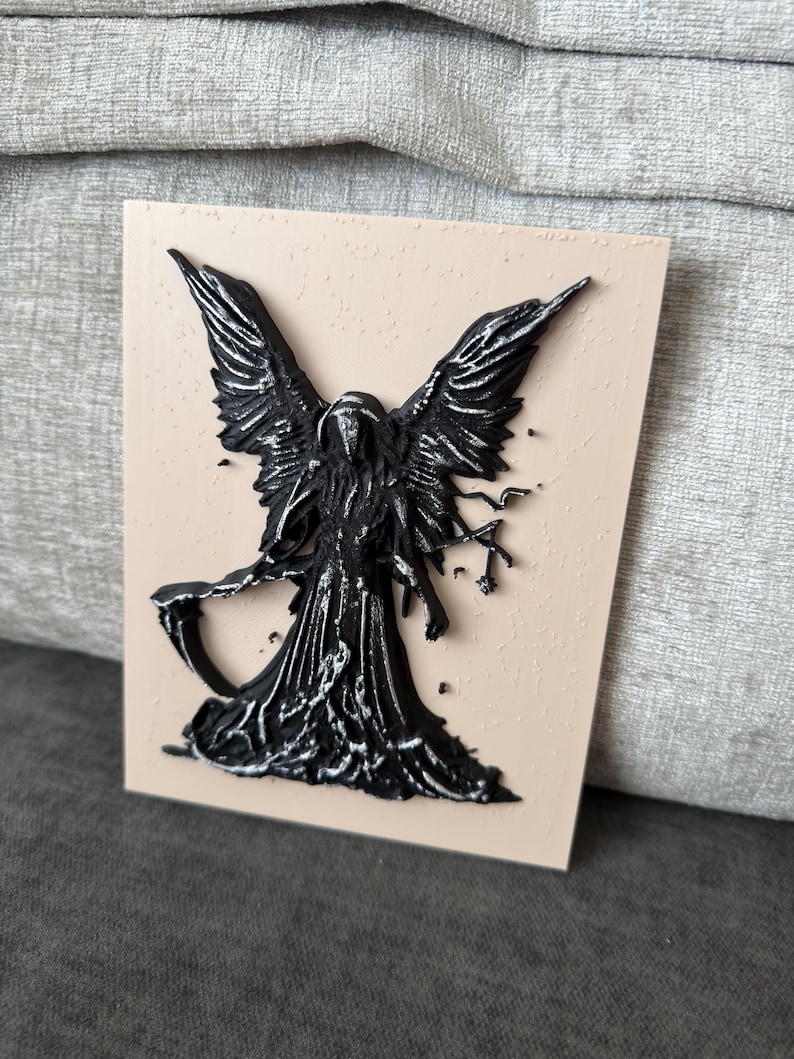 May include: A rectangular plaque with a light beige textured surface featuring a raised, three-dimensional black figure of a winged entity. The figure has large feathered wings, a hooded cloak, and holds a scythe in its right hand.