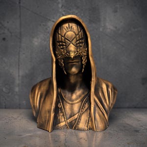 May include: A bronze-colored bust sculpture of a hooded figure wearing an ornate, filigree-style mask. The mask features intricate cut-out patterns across the eyes and nose, while the figure is draped in a hooded cloak with visible fabric folds.
