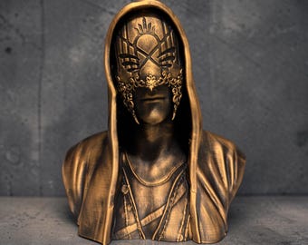 May include: A bronze-colored bust sculpture of a hooded figure wearing an ornate, filigree-style mask. The mask features intricate cut-out patterns across the eyes and nose, while the figure is draped in a hooded cloak with visible fabric folds.