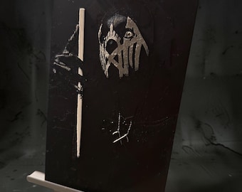 May include: A rectangular black plaque featuring the text SLEEP TOKEN and a central metallic silver graphic of a stylized mask. A thin vertical light-colored rod is positioned on the left side of the plaque, which rests on a small wooden easel.