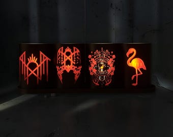 May include: A rectangular dark metal lamp featuring four distinct cut-out panels illuminated from within by a warm orange glow. The panels display intricate designs including geometric shapes, ornate floral patterns, and a flamingo silhouette.
