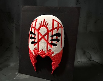May include: A white mask with a textured surface featuring red linear patterns and a red jagged lower section. The mask has three horizontal black cutouts on each side and is mounted on a flat black square panel with a matte finish.
