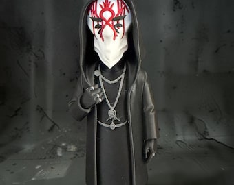 May include: A figurine wearing a long black hooded coat and matching boots. The figure has a white mask featuring intricate red markings. It is adorned with layered silver-toned necklaces and rings on its fingers, presenting a detailed, monochromatic aesthetic with bold red accents on the face.