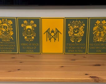 May include: A collection of eleven rectangular plaques arranged in a row. Ten plaques are dark green with gold-colored intricate illustrations and text, while the center plaque is yellow with a black geometric symbol and the text "THM".