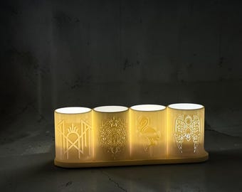 May include: A set of four cylindrical, off-white, translucent candle holders arranged on a matching rectangular base. Each holder features a unique, intricate cutout design, including a geometric emblem, a detailed crest, a flamingo, and wings.