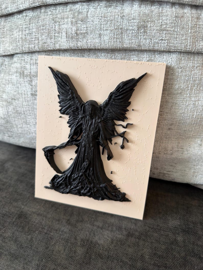 May include: A rectangular wall plaque featuring a raised, three-dimensional black figure of a winged entity with a hooded cloak. The figure holds a scythe in one hand and stands against a smooth, light beige background with a textured surface.