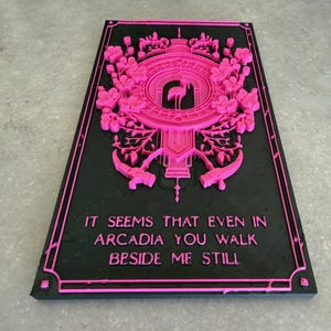 May include: A rectangular black plaque featuring a raised hot pink relief design. The top section displays a central flamingo silhouette surrounded by intricate floral patterns and geometric borders. The text reads: IT SEEMS THAT EVEN IN ARCADIA YOU WALK BESIDE ME STILL.