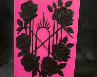 May include: A rectangular silicone mold in bright pink featuring a raised black design of five blooming roses arranged around a central arched gate structure with a pointed sunburst top. The mold has a smooth surface and deep relief details.