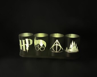 May include: A set of four cylindrical black candle holders arranged on a rectangular base. Each holder features a cutout design illuminated from within, displaying the letters HP, a face with round glasses, a triangle symbol, and a castle silhouette.