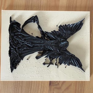 May include: A rectangular light beige plaque featuring a three-dimensional black relief sculpture of a winged figure with a flowing garment. The figure is positioned diagonally across the surface, displaying detailed textures on the wings and fabric.