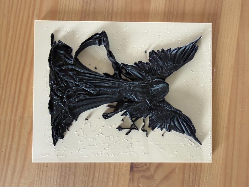 May include: A rectangular light beige plaque featuring a three-dimensional black relief sculpture of a winged figure with a flowing garment. The figure is positioned diagonally across the surface, displaying detailed textures on the wings and fabric.