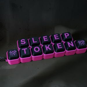 May include: A keychain featuring black square keys with pink lettering that spells out SLEEP TOKEN, arranged in two rows. The keys are set in a bright pink ridged base, and a small metal carabiner is attached to the left side of the structure.