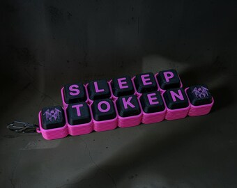 May include: A keychain featuring black square keys with pink lettering that spells out SLEEP TOKEN, arranged in two rows. The keys are set in a bright pink ridged base, and a small metal carabiner is attached to the left side of the structure.