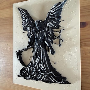 May include: A rectangular beige plaque featuring a raised, three-dimensional black figure of a winged entity holding a scythe. The figure has detailed, textured wings and a flowing robe, with small, scattered black paint droplets across the surface.
