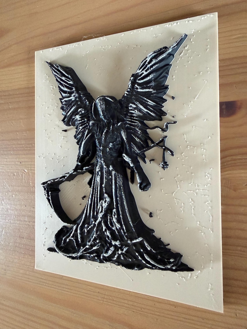 May include: A rectangular beige plaque featuring a raised, three-dimensional black figure of a winged entity holding a scythe. The figure has detailed, textured wings and a flowing robe, with small, scattered black paint droplets across the surface.