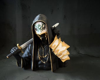 May include: A sculpted bust of a hooded figure wearing a white mask with green markings, a black robe with gold trim, and a gold chain necklace. The figure holds a silver sword over the shoulder and wears a white and gold armored gauntlet on one arm.