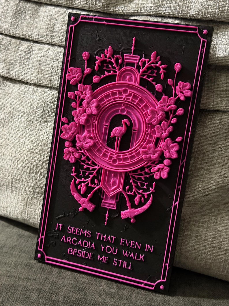 May include: A rectangular black plaque with raised hot pink relief details. The design features a central flamingo inside a circular frame, surrounded by floral motifs and two scythes. The text reads: IT SEEMS THAT EVEN IN ARCADIA YOU WALK BESIDE ME STILL.