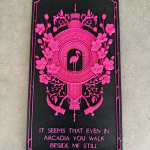 May include: A rectangular black plaque featuring a raised hot pink design of a flamingo centered within a circular frame, surrounded by floral motifs and two scythes. The text reads: IT SEEMS THAT EVEN IN ARCADIA YOU WALK BESIDE ME STILL.