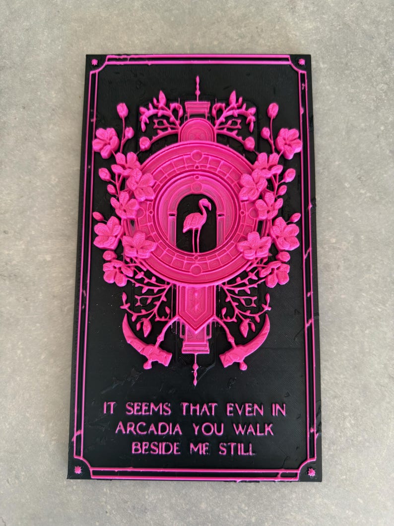 May include: A rectangular black plaque featuring a raised hot pink design of a flamingo centered within a circular frame, surrounded by floral motifs and two scythes. The text reads: IT SEEMS THAT EVEN IN ARCADIA YOU WALK BESIDE ME STILL.