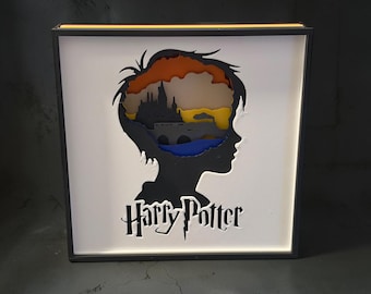 May include: A square shadow box display featuring a black silhouette of a person's head in profile. Inside the silhouette is a layered paper cutout of a castle and bridge against a sunset-colored background. The text Harry Potter is below.