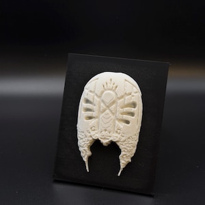 May include: An off-white, oval-shaped relief sculpture featuring intricate, symmetrical geometric carvings and textured, organic-looking edges. The piece is mounted on a flat, rectangular black backing, creating a high-contrast display.
