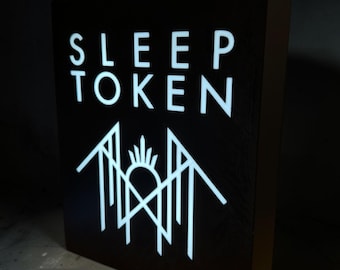 May include: A rectangular black light box featuring the white text SLEEP TOKEN at the top and a white geometric logo below. The sign is illuminated from within, creating a high-contrast display against the dark background of the box structure.