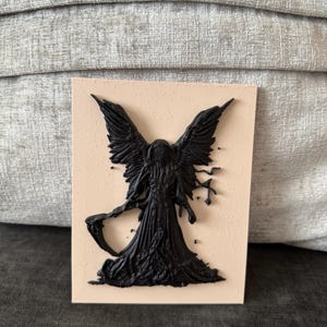 May include: A rectangular plaque featuring a raised, black, three-dimensional relief of a hooded figure with large wings and a scythe. The figure is centered on a smooth, light beige, flat surface, creating a stark contrast between the dark, textured silhouette and the pale background.