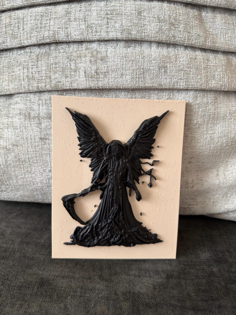 May include: A rectangular plaque featuring a raised, black, three-dimensional relief of a hooded figure with large wings and a scythe. The figure is centered on a smooth, light beige, flat surface, creating a stark contrast between the dark, textured silhouette and the pale background.