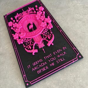 May include: A rectangular black plaque with raised hot pink relief details featuring a central flamingo silhouette, floral motifs, and the text: IT SEEMS THAT EVEN IN ARCADIA YOU WALK BESIDE ME STILL. The design is framed by a thin pink border.