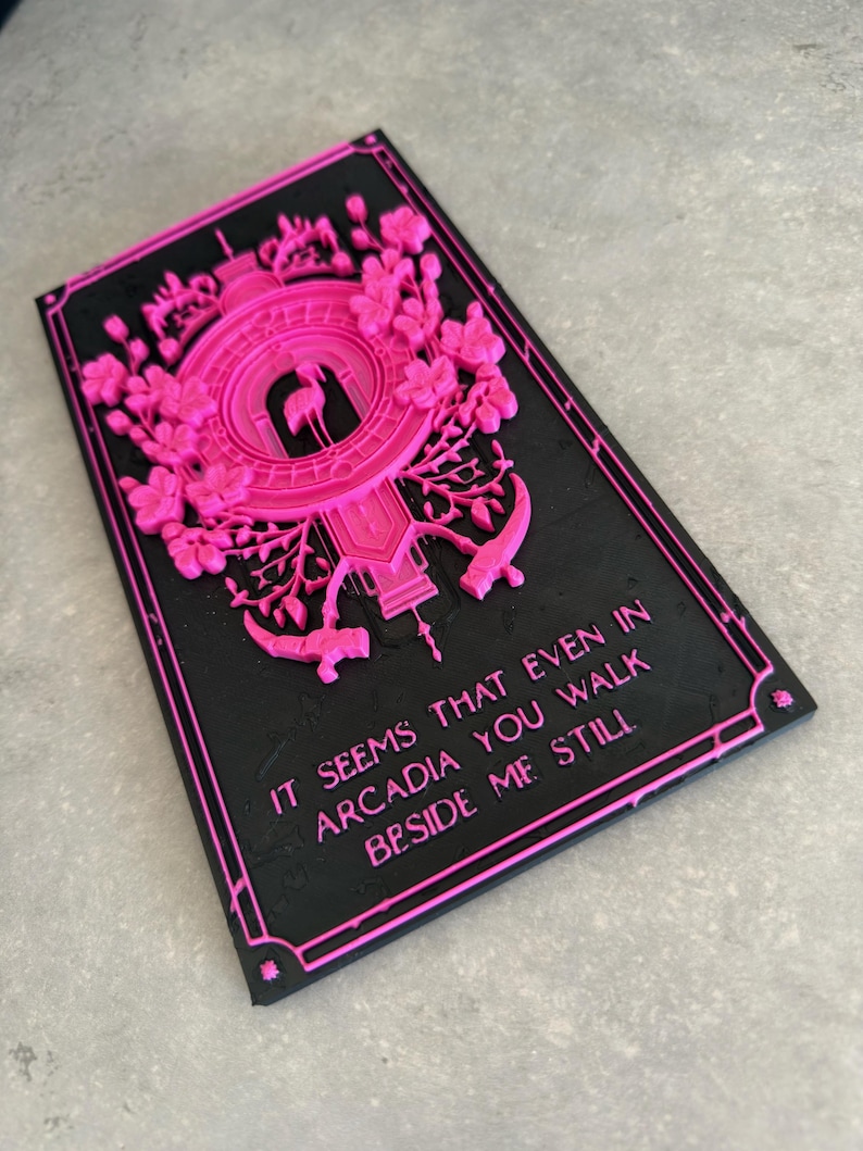 May include: A rectangular black plaque with raised hot pink relief details featuring a central flamingo silhouette, floral motifs, and the text: IT SEEMS THAT EVEN IN ARCADIA YOU WALK BESIDE ME STILL. The design is framed by a thin pink border.