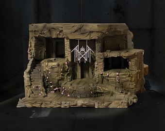 May include: A tan-colored, rocky diorama display base featuring a multi-level structure with carved stone stairs, textured walls, and small pink floral accents. A white, geometric, symmetrical emblem hangs from the center of the upper archway.