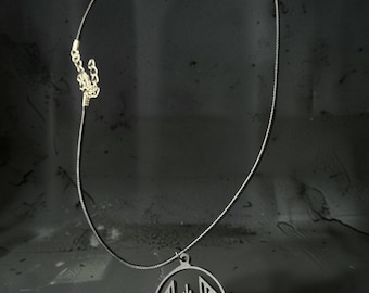 May include: A black cord necklace featuring a circular pendant with a geometric, angular cutout design. The pendant is dark gray and suspended from a thin, braided black cord that includes a gold-toned metal lobster clasp and an adjustable chain.