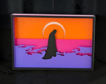May include: A rectangular light box featuring a layered paper art scene. The design displays a black silhouette of a cloaked figure standing before a large white circular sun against a vibrant orange sky, a pink horizon, and purple foreground waves.