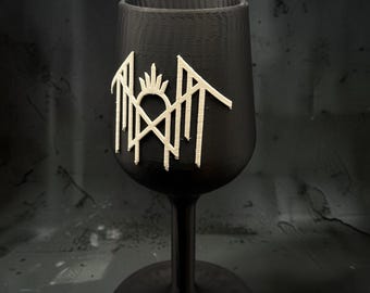 May include: A matte black stemmed goblet featuring a raised, off-white, angular, symmetrical emblem on the front. The emblem consists of sharp, pointed lines and a central crown-like shape. The goblet has a smooth, cylindrical bowl and base.