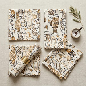 May include: A set of four cream-colored cloth napkins with a repeating pattern of brown fish illustrations. One napkin is rolled and held with a gold napkin ring. A small bowl of salt, a spoon, and a sprig of greenery are also present.