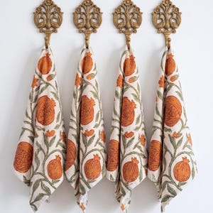 May include: Four decorative brass wall hooks with ornate floral designs hold matching cream-colored towels. Each towel features a repeating pattern of orange pomegranates and green leaves.