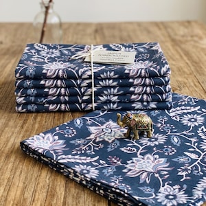 Op de afbeelding: Een stapel van vijf gevouwen marineblauwe katoenen servetten met een wit en lichtblauw bloemenpatroon, samengebonden met wit touw en een label met de tekst JAIPUR HANDCRAFTED Cotton Napkins Hand Block Printed, met een kleine koperen olifantfiguur.